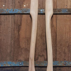 Op de afbeelding: Twee onafgewerkte houten bijlhandvatten, staand tegen een rustieke houten achtergrond. De lichtgekleurde handvatten hebben een glad, gebogen ontwerp. De achtergrond is een mix van bruine houten planken en een blauwe metalen strip.