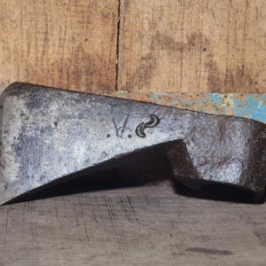 May include: A weathered, antique axe head with a dark, metallic finish. The axe head features a flat, beveled blade and a cylindrical socket for the handle. The surface shows signs of age and use, with visible markings. The axe head is resting on a wooden surface.
