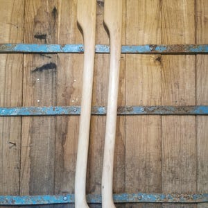 Könnte beinhalten: Zwei Ruder aus Holz mit hellem, natürlichem Finish. Die Ruder haben einen langen, schlanken Schaft und ein breites, flaches Blatt. Der Hintergrund ist eine Holzwand mit blauen Metallakzenten. Die Ruder sind wahrscheinlich für ein Boot.