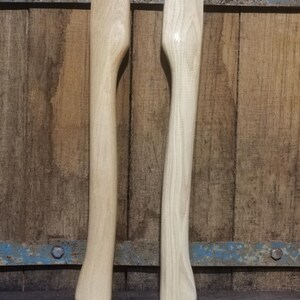 Op de afbeelding: Twee onafgewerkte houten bijlhandvatten. De lichtgekleurde handvatten hebben een glad, gebogen ontwerp om vast te pakken. De houtnerf is zichtbaar. De achtergrond is een verweerd houten oppervlak met een blauw metalen accent.