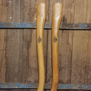 May include: Two wooden axe handles with a light brown finish. The handles are long and slender, with a curved design for a comfortable grip. The handles have a small hole at the bottom for attaching the axe head. The background is a wooden wall.