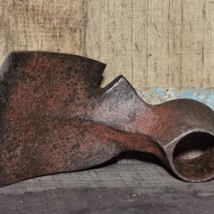 Op de afbeelding: Een verweerde, roestige metalen bijlkop met een gebogen blad en een cilindrisch oog voor de steel. De bijlkop is donkerroodbruin van kleur en vertoont tekenen van ouderdom en gebruik. De achtergrond is een houten oppervlak.