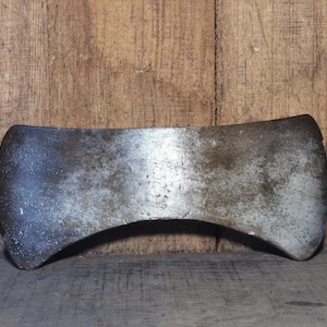 Peut inclure: Une tête de hache à double tranchant avec une lame incurvée. Le métal présente une apparence usée et vieillie, avec de la rouille et une décoloration visibles. La tête de hache est posée sur un fond en bois.