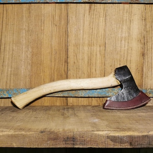 Puede incluir: Un hacha forjada a mano con mango de madera y funda de cuero. El hacha está apoyada en un estante de madera.
