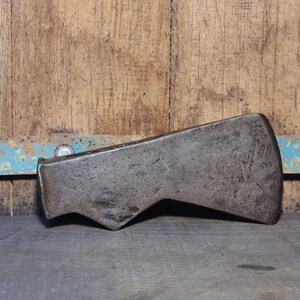Könnte beinhalten: Ein verwitterter, antiker Axtkopf mit dunklem, metallischem Finish. Die Axt hat eine markante, abgewinkelte Klinge und ein abgenutztes Aussehen, das auf Alter und Gebrauch hindeutet. Der Hintergrund ist eine rustikale Holzoberfläche.