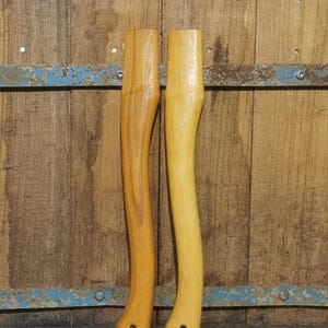 Op de afbeelding: Twee houten bijlhandvatten, één lichtbruin en één lichtgeel, worden getoond tegen een rustieke houten achtergrond. De handvatten hebben een gebogen ontwerp en een gat aan de basis voor het bevestigen van de bijlkop. De houtnerf is zichtbaar.