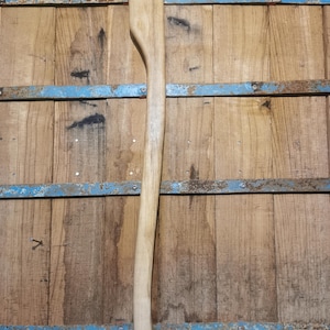 Op de afbeelding: Een nieuwe, onafgewerkte bijlsteel van lichtgekleurd hout. De steel is lang en slank, met een gebogen gedeelte om vast te pakken. De achtergrond is een houten oppervlak met blauwe metalen accenten.