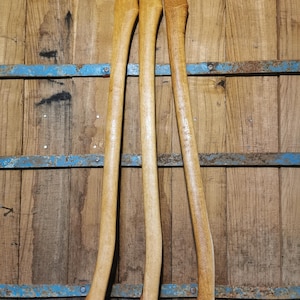 Könnte beinhalten: Drei Holzbeile mit hellbraunem Finish werden vor einem rustikalen Holzhintergrund präsentiert. Die Griffe sind lang und gebogen und haben ein glattes, poliertes Aussehen. Die Holzmaserung ist sichtbar und trägt zur natürlichen Ästhetik bei.