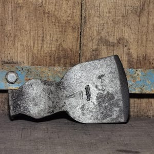 Op de afbeelding: Een verweerde, zilverkleurige bijlkop met een versleten uiterlijk. De bijlkop ligt op een houten oppervlak, met een blauwe en roestige metalen balk op de achtergrond. De bijlkop heeft een gestempeld logo.