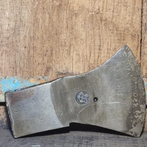 Peut inclure: Une tête de hache vintage avec une finition métallique usée. La tête de hache présente un emblème circulaire et un petit rivet sombre. Le fond est une surface en bois vieilli avec des traces de peinture bleue.