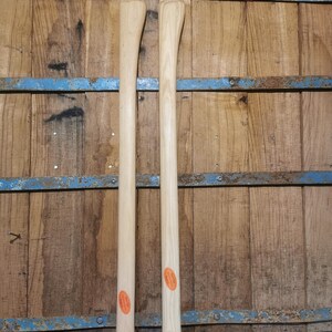 Puede incluir: Dos mangos de hacha de madera nuevos y sin terminar. Los mangos son de color claro con un acabado liso y una ligera curva. Cada mango tiene una pegatina naranja cerca de la base. El fondo es una pared de madera con detalles metálicos azules.