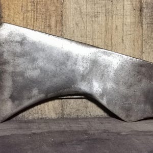 May include: A vintage, weathered axe head with a curved blade and a silver-gray finish. The axe head is set against a wooden background, showcasing its aged appearance and design. The axe head is a tool.