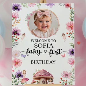 May include: A welcome sign for a first birthday celebration. The sign features a photo of a smiling baby girl, surrounded by floral and fairy illustrations. Text reads "Welcome to Sofia, fairy first birthday."