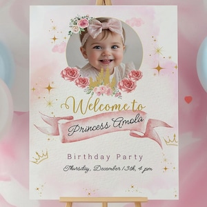 May include: A party sign on a wooden easel with the text "Welcome to Princess Amola" and a photo of a smiling baby. The sign is decorated with pink and gold accents, roses, and crowns. The party is on Thursday, December 13th, at 4 pm.