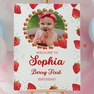 Puede incluir: Un letrero blanco sobre un caballete de madera con una foto de un bebé rodeado de fresas. El letrero dice "Welcome to Sophia Berry First Birthday!" en escritura roja. Ilustraciones de fresas rojas están dispersas alrededor del letrero.