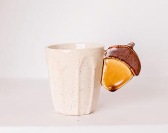 May include: Cream-colored ceramic mug with a faceted design. The handle is shaped like a brown and yellow acorn. The mug has a speckled texture and a glossy finish, suitable for hot beverages.