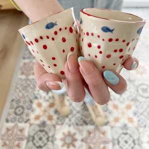 May include: Two white ceramic mugs with red polka dots and blue fish designs. The mugs have a slightly flared shape and are held in a person's hand.