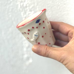 Könnte beinhalten: Eine kleine, weiße Keramiktasse mit rotem Rand und roten Tupfen. An der Seite ist ein kleiner, weißer Fisch mit blauen Details angebracht. Ein blaues Blatt ist in der Nähe des Randes gemalt. Die Tasse wird in einer Hand gehalten.