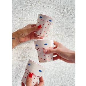 May include: Three white ceramic mugs with red polka dots and blue fish designs. The mugs are being held up in a toast.