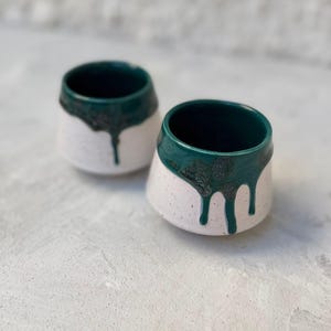 Può includere: Due tazze in ceramica fatte a mano con esterno bianco maculato e interno verde scuro. Lo smalto verde scuro cola sui lati, creando un design unico. Queste piccole tazze arrotondate sono perfette per caffè o tè.