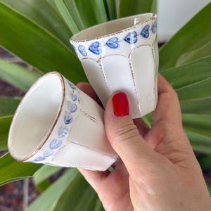 May include: Two small, off-white ceramic cups featuring a vertical paneled design and a hand-painted blue heart pattern around the upper rim. The edges of the cups and the vertical panels are accented with a distressed brown finish.