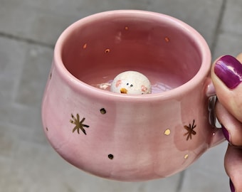 May include: A pink ceramic mug with a snowman figurine inside. The mug has gold star and dot accents. The snowman has a painted face. The mug is held by a person with purple nail polish.
