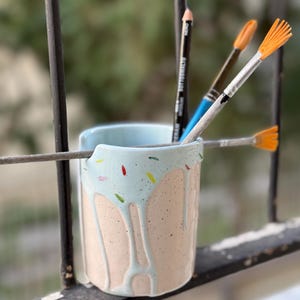Peut inclure: Un porte-crayons en céramique bleu clair avec un glaçage rose et blanc en forme de goutte et des pépites colorées. Le porte-crayons est rempli de pinceaux et d'un crayon.