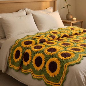 May include: A handmade crochet blanket featuring a sunflower design. The blanket has yellow petals, brown centers, and green and yellow edging. The blanket is draped over a bed with white pillows and a wooden headboard.