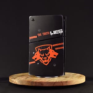 May include: A black PlayStation 5 console with a custom skin featuring an orange wolf head logo and the text "Call of Duty: The Truth Lies".