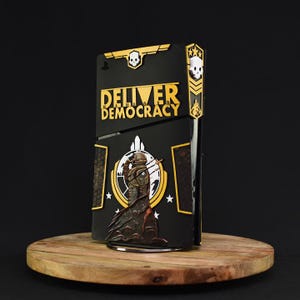 May include: Black and gold decorative console cover with the text "DELIVER DEMOCRACY". The cover features a soldier graphic and skull emblems. The console cover is displayed on a wooden surface.