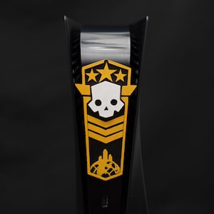 Pode incluir: Uma placa de console de videogame dourada e branca com um design de caveira e ossos cruzados. A placa tem três estrelas e uma insígnia de estilo militar.