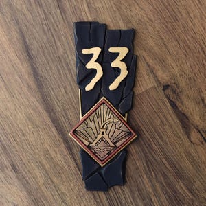 May include: A black and gold decorative plaque featuring the number 33 in gold. The plaque has a diamond-shaped emblem with a gold and red design. The plaque is on a wooden surface.