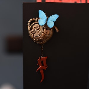 Può includere: Un ornamento a forma di cuore in stile steampunk con ingranaggi in bronzo e una farfalla azzurra. Un numero 17 rosso stilizzato è appeso sotto. L'ornamento è posto su uno sfondo nero.