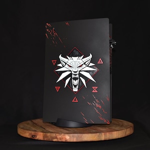 May include: Black console cover featuring a white wolf head emblem with red accents, set against a black background. The design includes red geometric shapes and splatter effects. The console cover is displayed on a wooden stand.