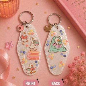 May include: Two decorative keychains with a white, speckled background. The front keychain features a cartoon goose reading books, while the back shows a goose under a blanket with a cup. Both have floral accents and gold keyrings.