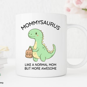 May include: White ceramic mug with a green dinosaur illustration holding a brown purse. The text "MOMMYSAURUS" is above the dinosaur and "LIKE A NORMAL MOM BUT MORE AWESOME" is below the dinosaur.