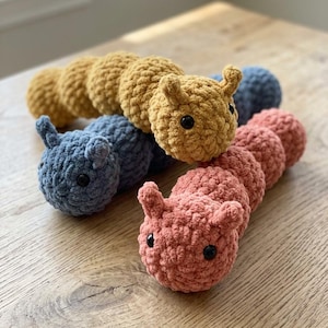 May include: Crocheted caterpillar toys in shades of yellow, blue, and pink. Each caterpillar has a rounded body and small antennae. The toys are arranged on a wooden surface.