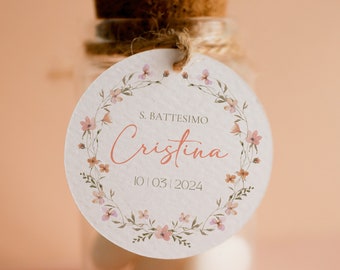 May include: A small glass jar with a cork lid and a white round tag tied with twine. The tag has a floral wreath design with the text "S. BATTESIMO Cristina 10.03.2024".