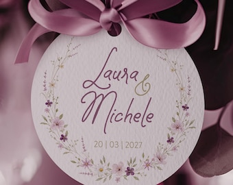 May include: A circular white tag featuring a floral wreath illustration in shades of purple and green. The text reads Laura & Michele with the date 20 | 03 | 2027. A mauve satin ribbon is tied in a bow at the top, with a long ribbon tail hanging down.