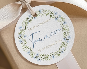 May include: A round white gift tag with a floral wreath of green leaves and light blue flowers. The text reads SANTA CRESIMA, Tommaso, and 10 OTTOBRE 2026. The tag is tied with a light beige satin ribbon around a tan gift box.