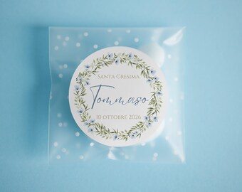 May include: A square, translucent white favor bag with a white polka dot pattern containing a round sticker. The sticker features a green leaf and blue flower wreath with the text SANTA CRESIMA, Tommaso, and 10 OTTOBRE 2026 printed in the center.