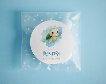 May include: A round white sticker with a watercolor illustration of a cute turtle, surrounded by blue watercolor accents. The sticker is inside a clear plastic bag with white polka dots. Text on the sticker reads "è nato Lorenzo 12 ottobre 2025".
