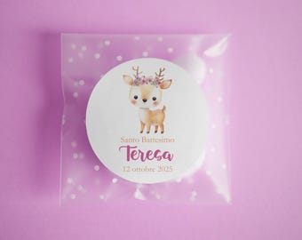 May include: A round white sticker with a watercolor illustration of a deer wearing a floral crown, inside a clear cellophane bag with white polka dots. The sticker has text that reads "Santo Battesimo Teresa 12 ottobre 2025". The background is a solid light purple.
