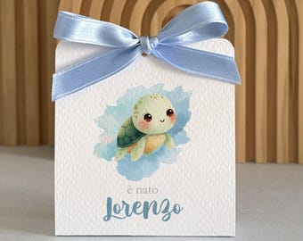 May include: A white rectangular card featuring a light blue satin ribbon bow at the top. The card displays a watercolor illustration of a small green sea turtle swimming in blue water, with the text "è nato Lorenzo" printed below.