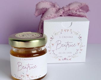 May include: A small glass jar of honey with a gold lid and a label that reads "Beatrice". A white box with a floral design and a pink ribbon bow is behind the jar. The box also has the name "Beatrice" on it.
