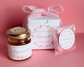 May include: A collection of personalized favors for a First Communion, including a small jar of honey, a gift box, and a wrapped treat. All items are decorated with floral accents and the name "Amanda" and the date "18 aprile 2026".