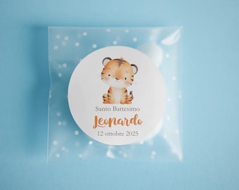 May include: A round sticker with a watercolor illustration of a tiger cub, with the text "Santo Battesimo Leonardo 12 ottobre 2025". The sticker is inside a clear plastic bag with white polka dots, set against a light blue background.