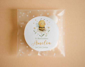 May include: A clear cellophane bag with white polka dots contains white candies. A round sticker with a cartoon bee on a flower is centered on the bag. The sticker has the text "S. Battesimo Amelia 24|05|2026".