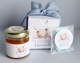 May include: A gift set featuring a jar of honey, a box, and a small wrapped item, all decorated with a sleeping teddy bear illustration. The box is tied with a light blue ribbon. The text "S. Battesimo Riccardo" is visible.