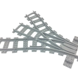 Puede incluir: Un desvío de vía de tren de juguete de plástico gris, diseñado para permitir que un tren cambie de dirección. La vía tiene una serie de raíles paralelos conectados por traviesas espaciadas uniformemente.